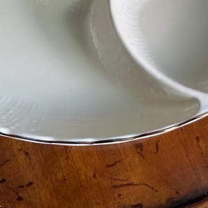 May include: A white ceramic divided serving dish with a scalloped edge and a metallic gold rim. The dish features a smooth, glossy finish and a subtle embossed pattern along the curved border, separating the interior into distinct sections.