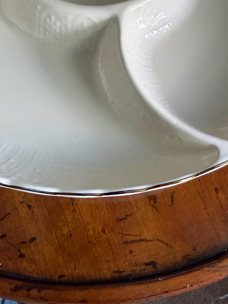 May include: A white ceramic divided serving dish with a scalloped edge and a metallic gold rim. The dish features a smooth, glossy finish and a subtle embossed pattern along the curved border, separating the interior into distinct sections.