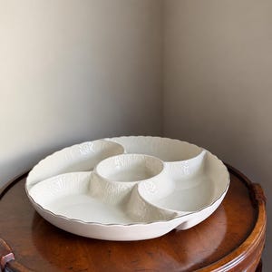 May include: A round, off-white ceramic serving platter featuring four distinct outer compartments and a central circular bowl. The dish has a scalloped edge and a subtle, embossed vertical pattern along the interior walls of each section.
