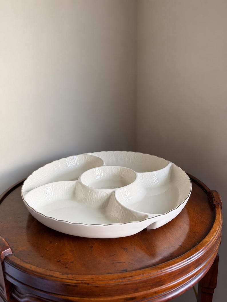 May include: A round, off-white ceramic serving platter featuring four distinct outer compartments and a central circular bowl. The dish has a scalloped edge and a subtle, embossed vertical pattern along the interior walls of each section.
