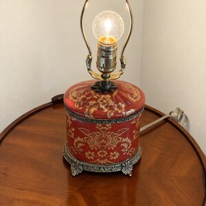 Może przedstawiać: Lampa stołowa z cylindryczną czerwoną podstawą ozdobioną złotym kwiatowym wzorem adamaszku. Lampa posiada ozdobną metalową podstawę z dekoracyjnymi n&oacute;żkami, mosiężną oprawkę i harfę, zwieńczoną pojedynczą świecącą żar&oacute;wką.
