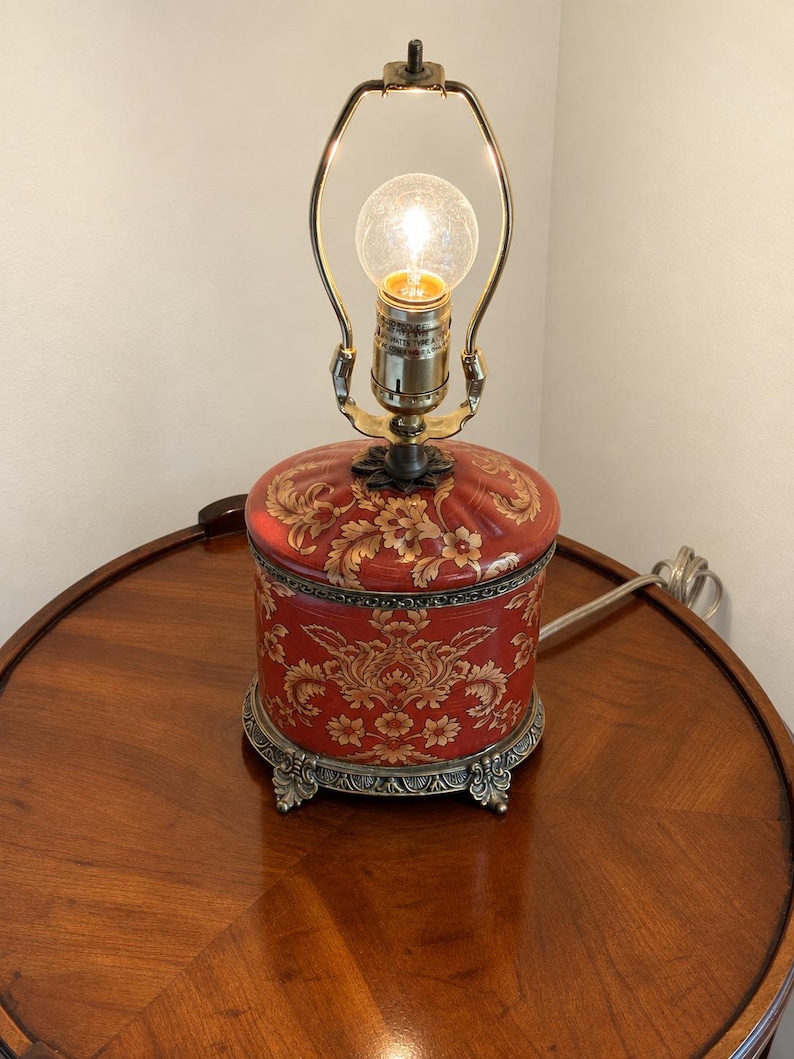 Może przedstawiać: Lampa stołowa z cylindryczną czerwoną podstawą ozdobioną złotym kwiatowym wzorem adamaszku. Lampa posiada ozdobną metalową podstawę z dekoracyjnymi n&oacute;żkami, mosiężną oprawkę i harfę, zwieńczoną pojedynczą świecącą żar&oacute;wką.