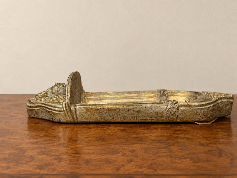 May include: A rectangular, boat-shaped decorative tray with a weathered, distressed gold and silver finish. The tray features a raised vertical divider at one end and ornate, sculpted relief details along the edges and the front section.