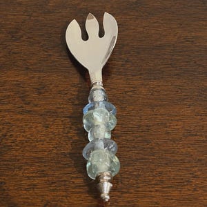 May include: A silver-toned metal serving fork featuring a three-pronged head and a decorative handle. The handle is composed of several stacked, translucent glass beads in shades of light blue, seafoam green, and clear, finished with a metal end cap.