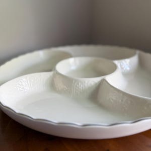 May include: A white ceramic serving dish with a scalloped edge and a central circular compartment surrounded by three larger sections. The exterior walls of the compartments feature a subtle, embossed vertical pattern with floral motifs.