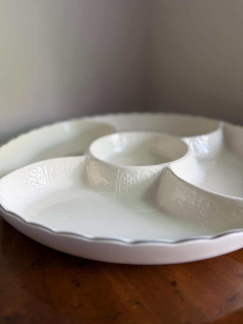 May include: A white ceramic serving dish with a scalloped edge and a central circular compartment surrounded by three larger sections. The exterior walls of the compartments feature a subtle, embossed vertical pattern with floral motifs.