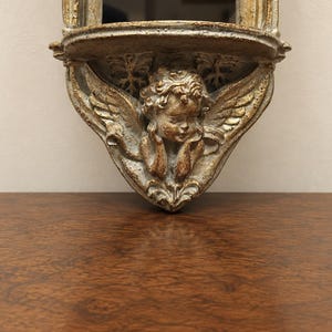 May include: A wall-mounted shelf featuring a sculpted cherub with wings and curly hair. The piece has a distressed finish with gold and grey tones, showcasing intricate relief details of foliage and a small platform for display purposes.