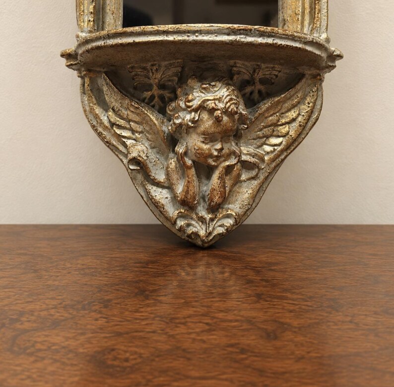 May include: A wall-mounted shelf featuring a sculpted cherub with wings and curly hair. The piece has a distressed finish with gold and grey tones, showcasing intricate relief details of foliage and a small platform for display purposes.