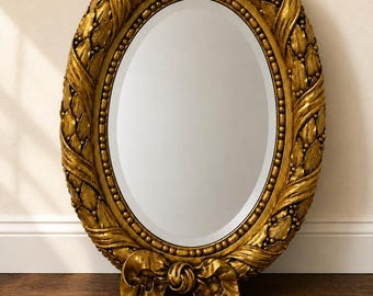 May include: An oval mirror featuring a thick, ornate gold frame decorated with sculpted leaf patterns, a beaded inner border, and a prominent bow detail at the base. The reflective glass surface is smooth and clear, set within the textured gold structure.