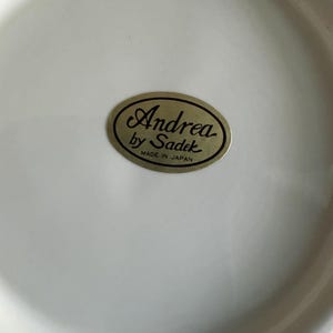 May include: A smooth, off-white ceramic surface featuring an oval-shaped gold sticker in the center. The sticker displays the black text Andrea by Sadek with the words MADE IN JAPAN printed in smaller black capital letters underneath the brand name.