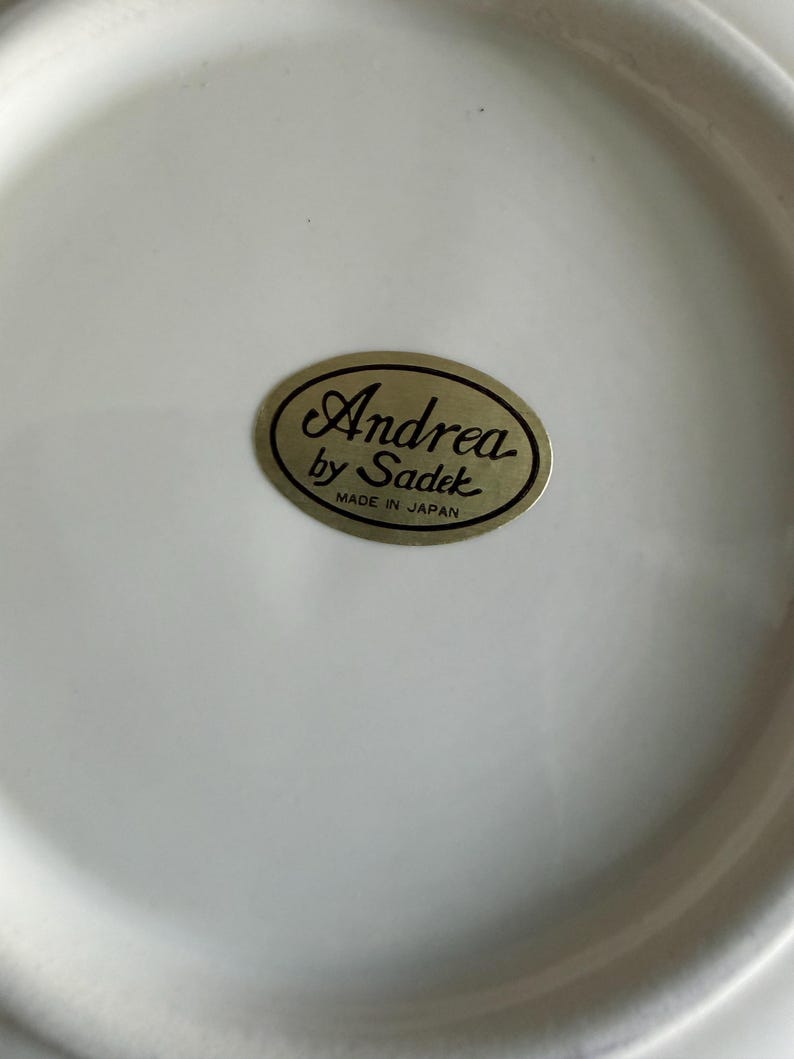 May include: A smooth, off-white ceramic surface featuring an oval-shaped gold sticker in the center. The sticker displays the black text Andrea by Sadek with the words MADE IN JAPAN printed in smaller black capital letters underneath the brand name.