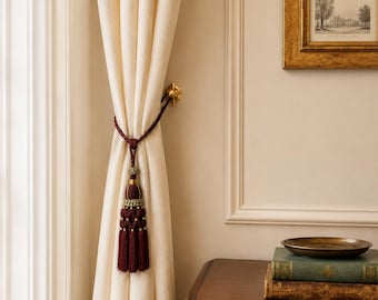 May include: A floor-length cream-colored curtain held back by a deep burgundy rope tieback featuring a decorative tassel with gold accents. The curtain is secured to a brass wall-mounted hook, creating a draped effect against the wall.