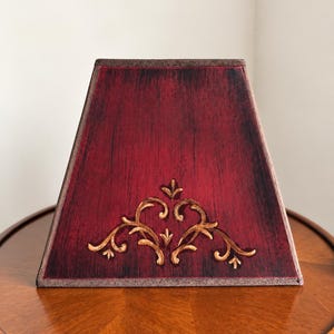May include: A trapezoidal lamp shade featuring a deep red finish with black distressed brush strokes. The front surface displays a raised, gold-toned scrollwork design centered near the bottom edge. The shade has a dark brown fabric trim along the top and bottom edges.
