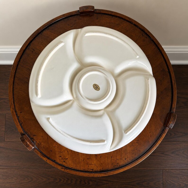 May include: A circular off-white ceramic serving platter featuring a central circular compartment surrounded by four curved, fan-shaped sections. The underside of the platter displays a small, oval-shaped gold sticker in the center.