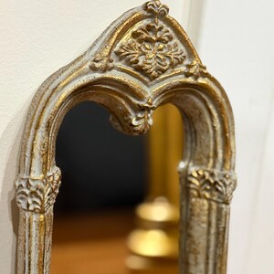 May include: An arched wall mirror featuring a distressed white and gold finish. The frame displays intricate relief carvings, including a prominent floral motif at the apex and decorative column-like details along the vertical sides of the mirror.