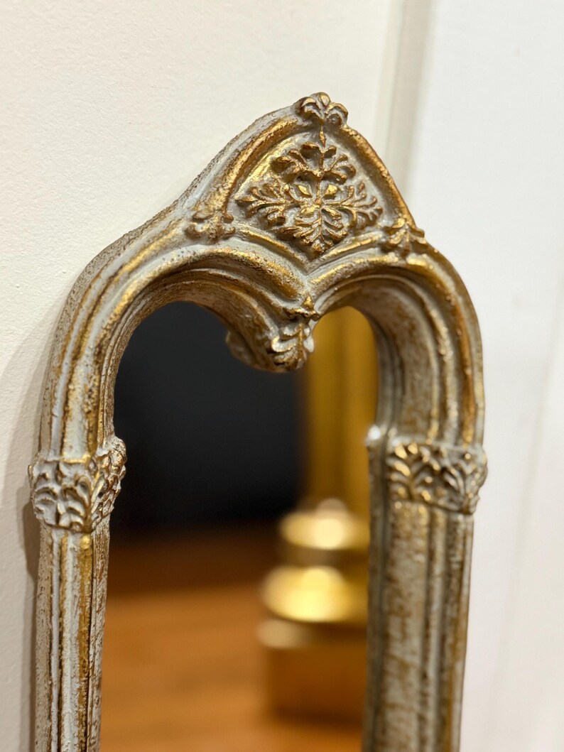 May include: An arched wall mirror featuring a distressed white and gold finish. The frame displays intricate relief carvings, including a prominent floral motif at the apex and decorative column-like details along the vertical sides of the mirror.
