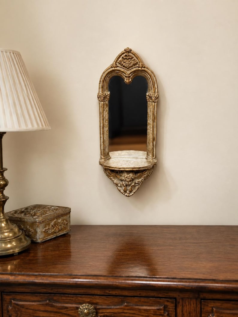 May include: A wall-mounted mirror featuring an arched gold frame with intricate carved detailing, including a small shelf at the base supported by a sculpted cherub figure. The frame has a distressed finish with visible gold and cream tones.