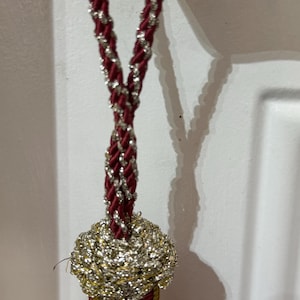 May include: A decorative tassel featuring a twisted cord in deep red and metallic silver. The top section is a rounded, woven knot in shimmering silver thread, transitioning into a cylindrical base wrapped in dark red cord with gold accents.