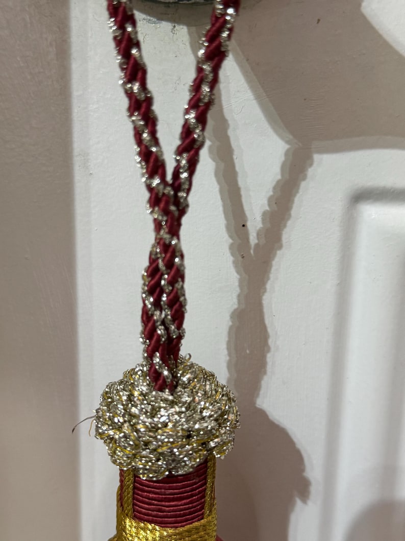 May include: A decorative tassel featuring a twisted cord in deep red and metallic silver. The top section is a rounded, woven knot in shimmering silver thread, transitioning into a cylindrical base wrapped in dark red cord with gold accents.
