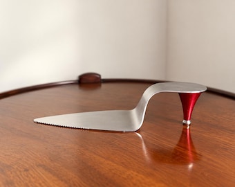 May include: A silver-colored metal cake server with a triangular blade and a serrated edge. The handle is shaped like a high-heeled shoe, featuring a metallic red conical heel support that elevates the back of the server from the surface.