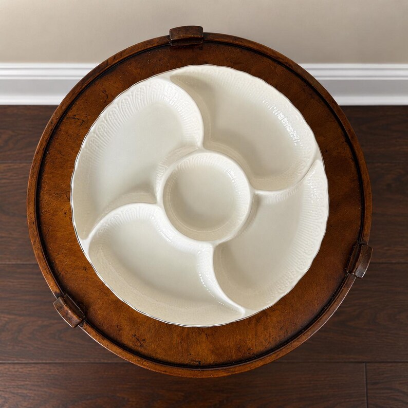 May include: A round, off-white ceramic serving platter featuring a central circular compartment surrounded by four distinct, curved sections. The outer rim of the platter is decorated with a subtle, embossed fan-like pattern along the edges.