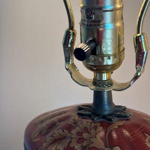 Może przedstawiać: Lampa stołowa z czerwoną podstawą w złoty wz&oacute;r kwiatowy oraz metalową oprawką w kolorze złota. Oprawka posiada czarny przełącznik pokrętłowy oraz etykietę UL z napisem E184994, PORTABLE LUMINAIRE oraz ISSUE NO. AK-49,307.