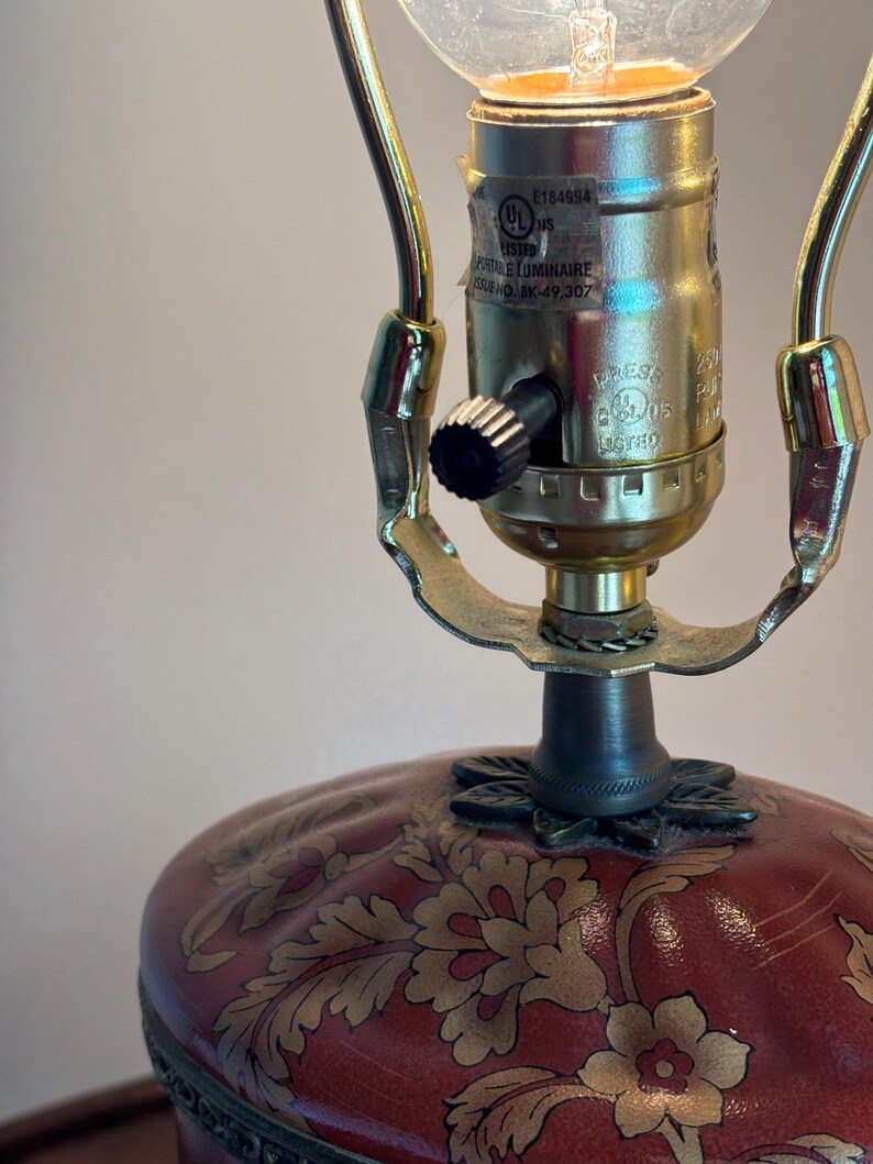 Może przedstawiać: Lampa stołowa z czerwoną podstawą w złoty wz&oacute;r kwiatowy oraz metalową oprawką w kolorze złota. Oprawka posiada czarny przełącznik pokrętłowy oraz etykietę UL z napisem E184994, PORTABLE LUMINAIRE oraz ISSUE NO. AK-49,307.