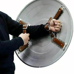 May include: A silver-colored, circular shield with a starburst design. The shield has brown leather straps and metal brackets for securing the shield to the arm. The shield is held by a person.