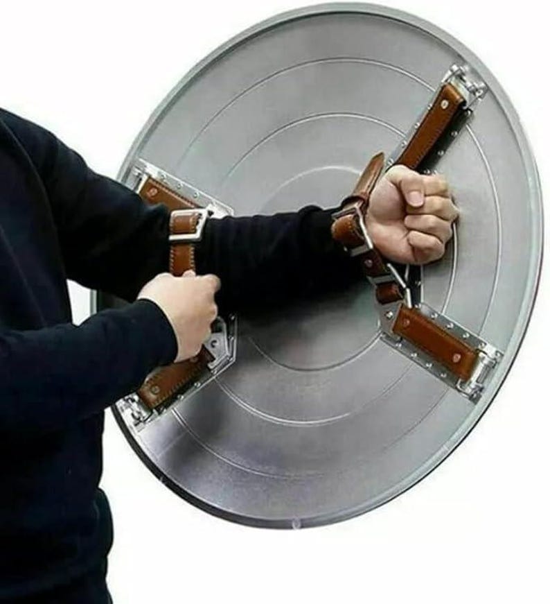 May include: A silver-colored, circular shield with a starburst design. The shield has brown leather straps and metal brackets for securing the shield to the arm. The shield is held by a person.