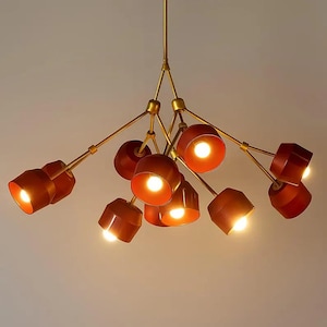 Peut inclure: Un lustre moderne avec une finition en laiton et de multiples abat-jours géométriques rouge-brun. Chaque abat-jour abrite une lumière vive, créant une lueur chaleureuse et accueillante. Le luminaire est suspendu au plafond.