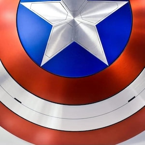 May include: A close-up of a Captain America shield. The shield features a central blue circle with a silver star, surrounded by a red ring, and a white stripe. The shield has a metallic finish.