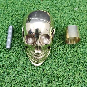 Pode incluir: Um objeto em forma de caveira dourada com uma peça cilíndrica correspondente e uma haste roscada prateada. A caveira tem traços faciais detalhados e está sobre uma superfície verde. A peça cilíndrica tem aproximadamente 3,8 cm de altura.