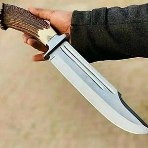 Peut inclure: Un grand couteau de chasse avec une lame en acier poli et un manche en bois de cerf. La poign&eacute;e est de couleur marron et cr&egrave;me naturelle, et la lame est tenue dans une main.