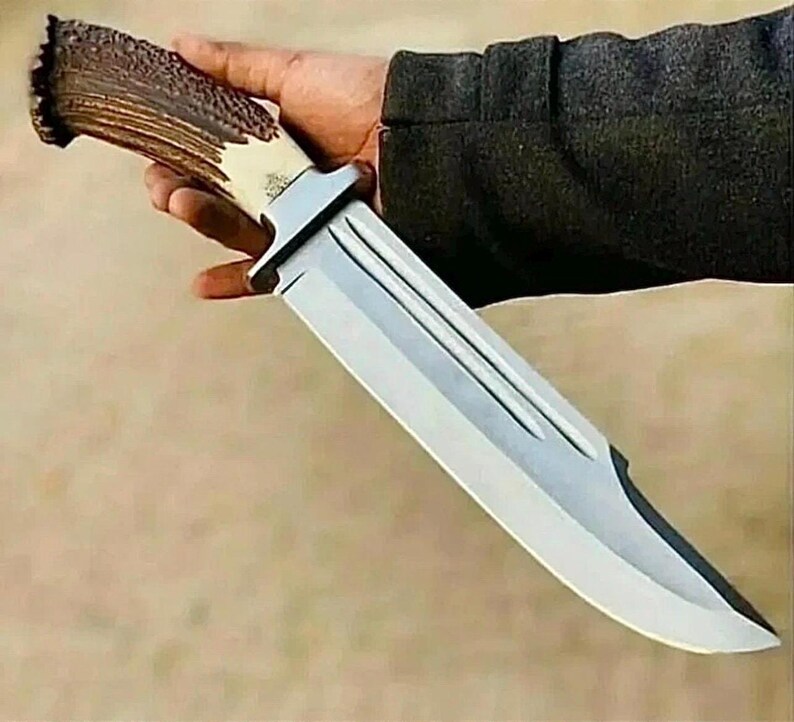 Peut inclure: Un grand couteau de chasse avec une lame en acier poli et un manche en bois de cerf. La poign&eacute;e est de couleur marron et cr&egrave;me naturelle, et la lame est tenue dans une main.