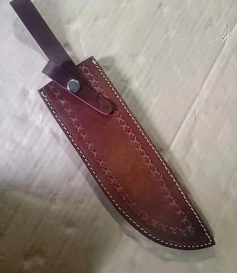 Peut inclure: Un &eacute;tui &agrave; couteau en cuir marron avec des coutures blanches et un motif d&eacute;coratif en forme d'&eacute;toile. L'&eacute;tui a une sangle avec une fermeture &agrave; bouton-pression en m&eacute;tal. L'&eacute;tui &agrave; couteau est con&ccedil;u pour prot&eacute;ger la lame.