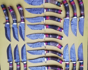 May include: A collection of Damascus steel knives with patterned blades and antler handles. The handles have red, blue, and green details. The knives are displayed on a yellow surface, highlighting their detailed designs and craftsmanship.