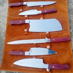 May include: A set of six stainless steel knives with burgundy handles, displayed in a brown leather knife roll. The blades are shiny and the handles have a dark red hue. The roll has a strap for carrying.