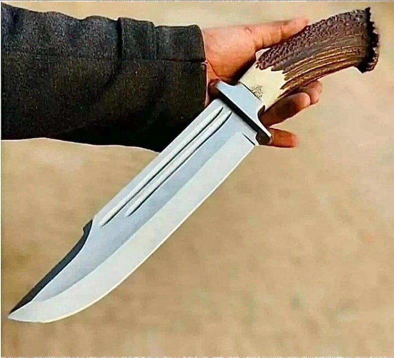 Peut inclure: Un grand couteau de chasse avec une lame argent&eacute;e polie et un manche en corne de cerf. La lame a un tranchant et une rainure d&eacute;corative. Le manche est un m&eacute;lange de marron clair et fonc&eacute;.