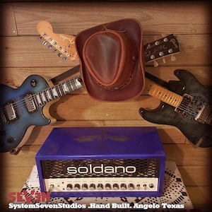 May include: A blue Soldano amplifier with a white front panel and numerous knobs and switches. A brown leather cowboy hat and several guitars are mounted on a wooden wall. The text "Soldano" is visible on the amplifier.