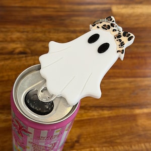 May include: A white ghost-shaped beverage can opener with two black oval eyes and a leopard print bow attached to the top. The opener is positioned on the silver tab of a pink aluminum soda can, featuring a flat, smooth surface design.