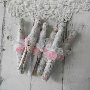 May include: A set of six wooden clothes pegs decorated with white paint, lace, and pink fabric flowers. The clothes pegs are arranged in a cluster on a white wooden surface.