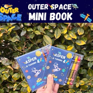 May include: Two small rectangular booklets with dark blue covers featuring space-themed graphics, including an astronaut, planets, and the text "ASPEN IS TURNING 4". Each booklet is packaged in a clear plastic sleeve with three crayons in purple, yellow, and red.
