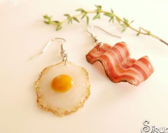 May include: A pair of earrings featuring a fried egg and a slice of bacon. The earrings are made of polymer clay and have silver hooks.