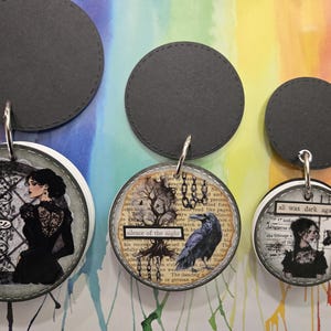 Puede incluir: Tres colgantes redondos decorativos con respaldo gris oscuro y aros plateados. Los colgantes presentan imágenes de temática gótica de mujeres, cuervos y texto. Un colgante dice "Elegance", otro "silence of the night" y el último "all was dark again."