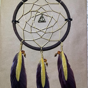 May include: A brown dreamcatcher with a circular frame wrapped in dark brown material. The web is made of thin, light-colored sticks. Three feathers hang below, each with a yellow center and dark purple edges. A gold-colored charm with a triangle and a line is in the center.