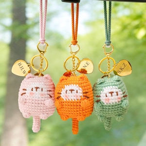 May include: Three crocheted cat keychains in pink, orange, and green, each with a gold-colored heart-shaped tag. The tags are engraved with initials. The keychains are hanging against a blurred green background.