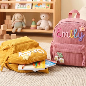 May include: A pink corduroy backpack with the name Emily embroidered in multicolored yarn and a small flower applique, alongside a yellow corduroy backpack with a white name applique and flower, both featuring zippered compartments.
