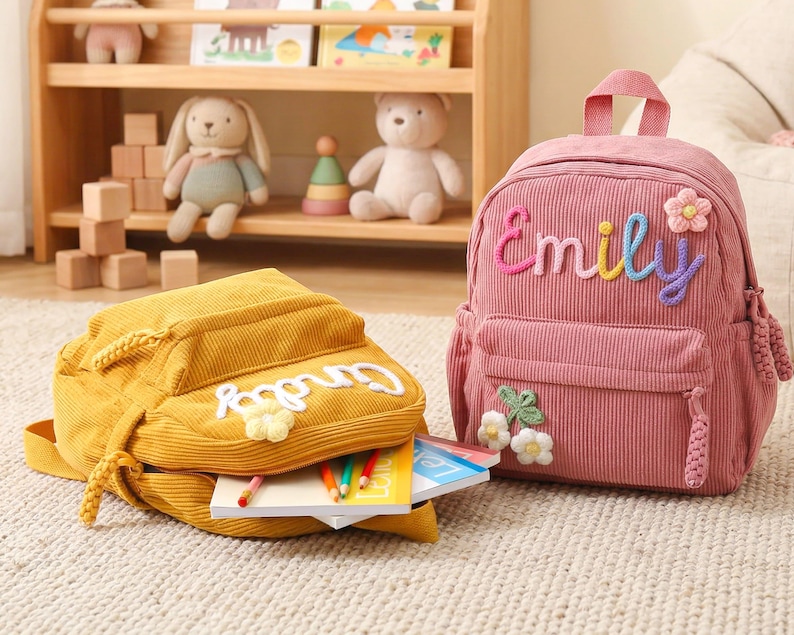 May include: A pink corduroy backpack with the name Emily embroidered in multicolored yarn and a small flower applique, alongside a yellow corduroy backpack with a white name applique and flower, both featuring zippered compartments.