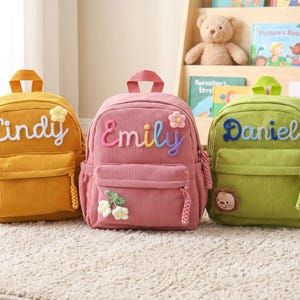 May include: Three corduroy backpacks in mustard yellow, dusty pink, and lime green. Each bag features a top handle, a front zippered pocket, and a personalized name embroidered in script: Cindy, Emily, and Daniel, with small floral or animal patches.