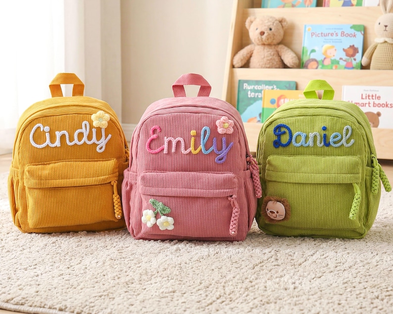 May include: Three corduroy backpacks in mustard yellow, dusty pink, and lime green. Each bag features a top handle, a front zippered pocket, and a personalized name embroidered in script: Cindy, Emily, and Daniel, with small floral or animal patches.